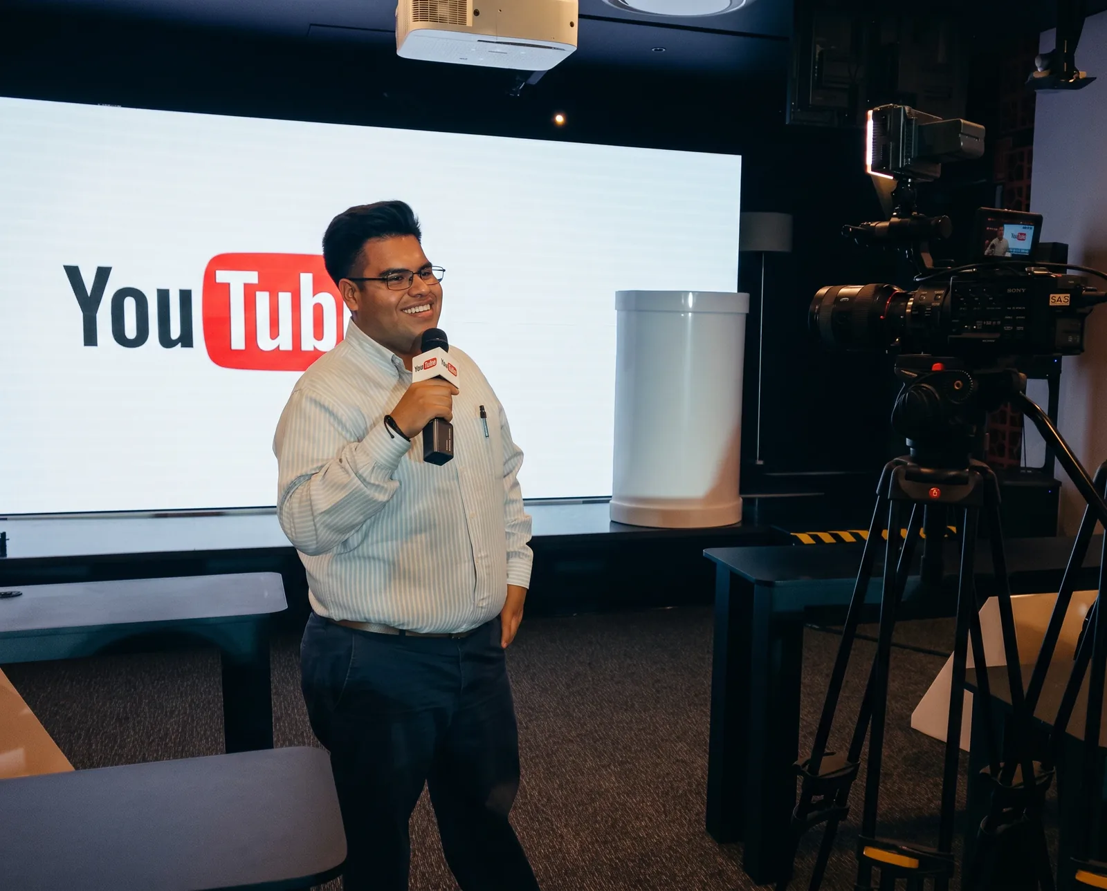 John Chávez dando una conferencia en un escenario de YouTube, con micrófono y pantalla del logo detrás