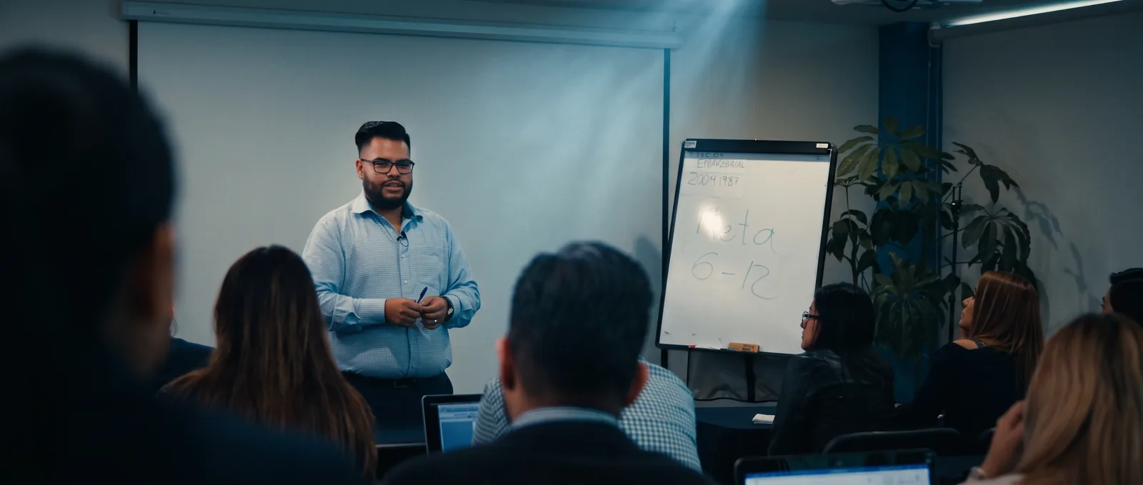 John Chávez imparte una clase en un salón de formación; los alumnos lo escuchan en primer plano y tras él se lee «Meta 6-12» en un rotafolio.